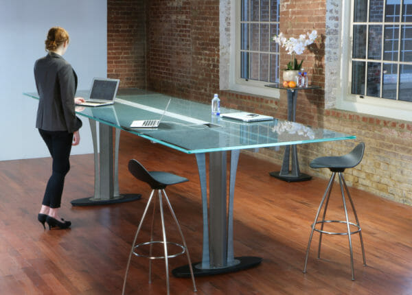 A woman stands at a Stoneline Designs Custom Tangent Conference Table. The table was designed with a stand-up height and incorporates glass and metal detailing and integrated wiring.